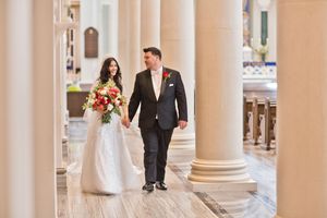 Bride and groom walk hand-in-hand through elegant church colonnade, bride holding vibrant red and orange bouquet.
