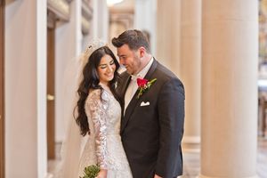 Bride and groom share a romantic moment among elegant columns, bride in lace gown with tiara, groom in dark suit with red boutonniere.