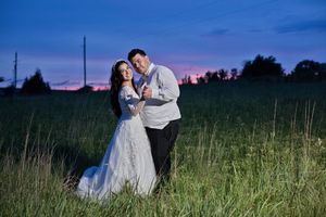 Couple in wedding attire embracing in a green field at twilight with a stunning purple and blue sunset sky.