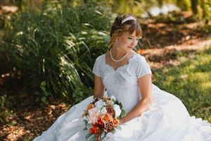 Bride in white gown holding orange and pink bouquet, seated outdoors near lush greenery on her wedding day.