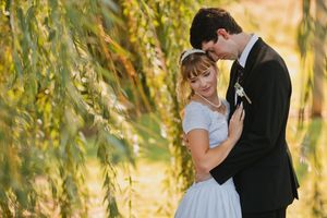 Bride and groom embrace under weeping willow tree in golden sunlight during outdoor wedding photo session.