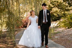 Bride and groom walking hand in hand on a garden path under weeping willow trees on their wedding day.