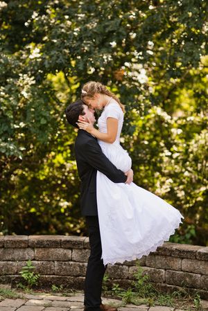 Groom lifts bride in white wedding dress for a romantic kiss outdoors, surrounded by lush green trees.