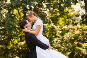 Groom lifting bride in white dress sharing a kiss outdoors with lush green trees in the background.