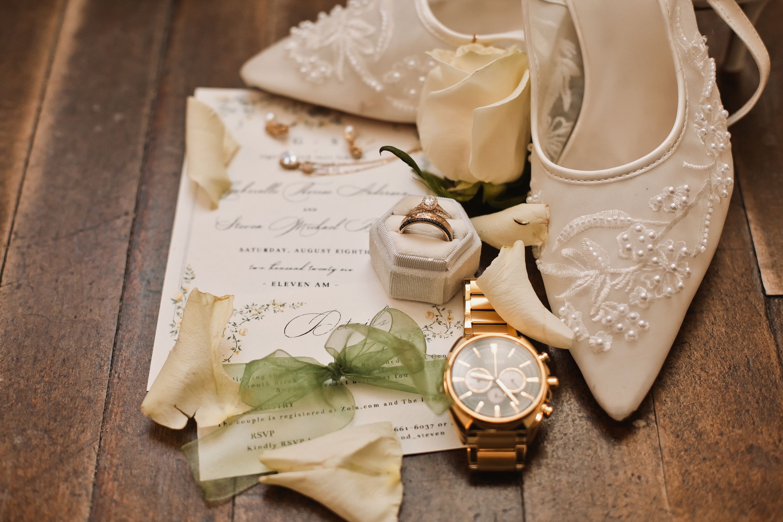 Elegant wedding flat lay with lace heels, gold watch, ring box, floral invitation, and a white rose on wood.