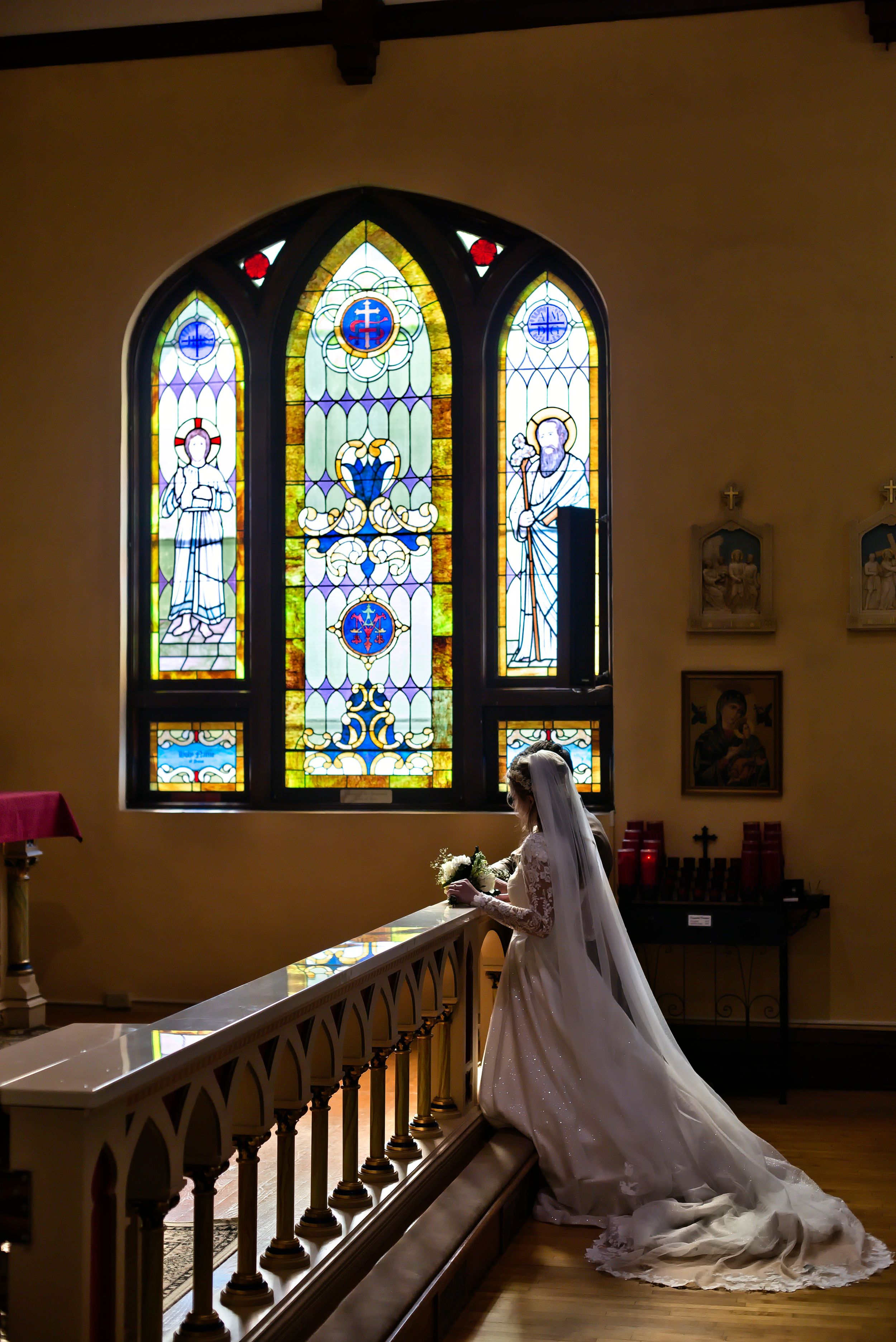 Bride in white gown and veil stands by church altar rail before colorful stained glass windows on her wedding day.