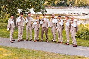Groomsmen in tan suits pose humorously on a scenic riverside path, adjusting their ties and collars for a wedding photo.