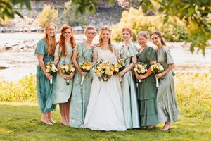 Bride in white gown with six bridesmaids in mismatched sage green dresses holding yellow bouquets outdoors.