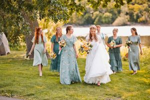 Bride in white gown walks with bridesmaids in sage green dresses holding yellow bouquets outdoors near a lake.