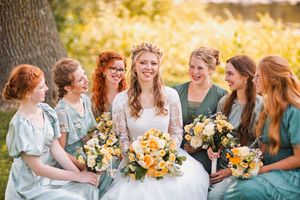 Bride and bridesmaids in sage green dresses laughing together outdoors holding yellow and white bouquets.