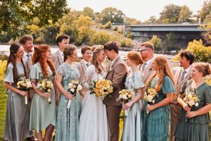 Wedding party poses outdoors, bridesmaids in sage green dresses holding yellow bouquets, as bride and groom share a kiss.