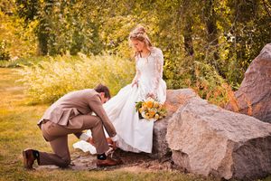 Groom kneeling to fix bride's shoe outdoors among rocks, bride holding yellow bouquet in white lace dress, fall wedding.