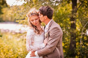 A bride and groom embrace outdoors, she wears a floral crown and lace dress, he wears a tan suit with boutonniere.