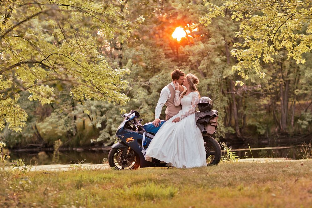 Newlywed couple kissing on a motorcycle at sunset surrounded by lush green trees in a romantic outdoor setting.