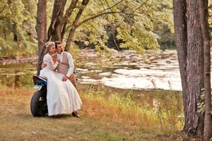 Pregnant couple sitting on a motorcycle near a river surrounded by trees in a romantic outdoor maternity photo shoot.
