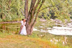 Bride and groom sharing a romantic kiss under a large tree by a peaceful river on their wedding day.
