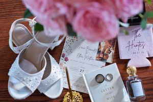 Elegant wedding flat lay with white bridal heels, pink roses, rings, perfume, invitation, and The Hunts koozies on wood.
