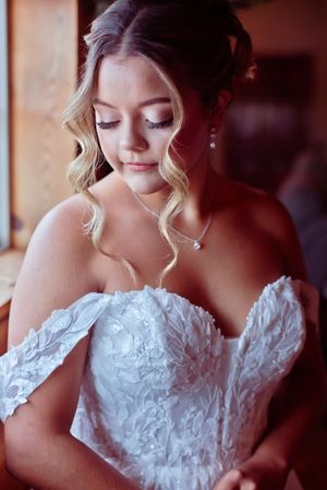 Bride in off-shoulder lace wedding gown with pearl necklace, looking down elegantly with soft curls and bridal makeup.
