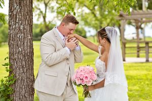 Groom in tan suit kissing bride's hand outdoors, bride holding pink bouquet in white wedding dress and veil.