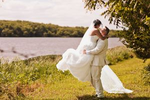 Groom lifts bride in white wedding gown for a romantic kiss by a scenic lakeside on their wedding day.