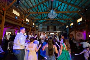 Guests dancing together at a rustic barn wedding reception with chandelier lighting and colorful purple uplighting.