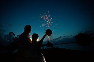 Silhouette of a bride and groom holding a bouquet toward fireworks bursting in a twilight sky over water.