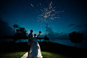 Bride and groom silhouette watching fireworks explode over waterfront at twilight during romantic outdoor wedding celebration.