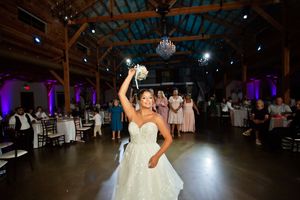 Bride in strapless ball gown tosses bouquet to guests during wedding reception in rustic barn venue.