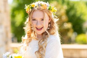 Smiling blonde bride wearing floral crown with yellow and white flowers, holding bouquet outdoors.