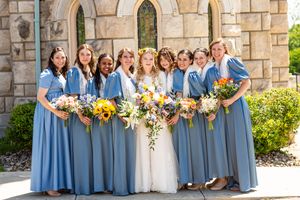 Bride and bridesmaids in blue dresses holding colorful bouquets with sunflowers outside a stone church.
