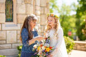 Bride in floral crown holds bouquet while older woman adjusts ring outside stone church on sunny wedding day.