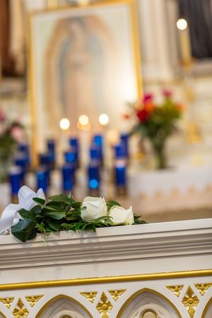 White roses rest on a ornate church altar with blue candles and Our Lady of Guadalupe image in soft focus background.