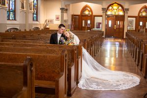 Bride and groom share an intimate moment in a wooden church pew, with a long flowing white veil trailing down the aisle.