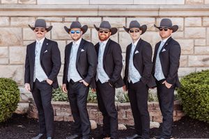 Five groomsmen in black suits, cowboy hats, and sunglasses pose outdoors against a stone wall at a western-themed wedding.