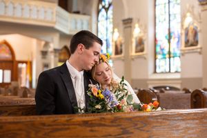 Bride and groom sitting closely together in church pew with colorful floral bouquet, stained glass windows in background.