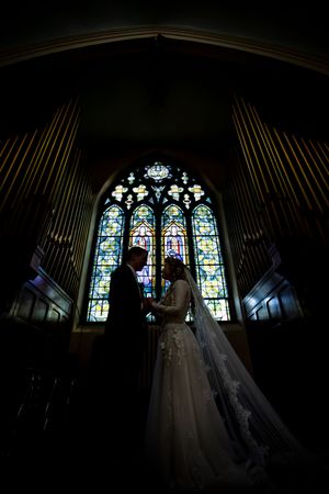 Silhouette of bride and groom holding hands before a stunning stained glass church window.