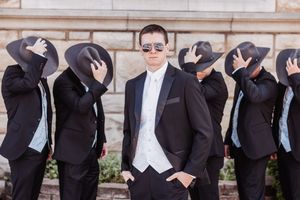 Groom in black tuxedo poses confidently with groomsmen tipping fedora hats in unison outdoors.