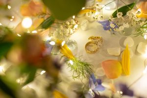 Gold wedding rings rest among colorful flower petals and twinkling fairy lights in a romantic floral arrangement.