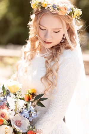 Blonde bride in white lace dress holding colorful bouquet, wearing floral crown of yellow and blue flowers.