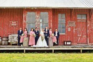 Wedding party posing in pink and navy attire before a rustic red barn venue on a sunny day.