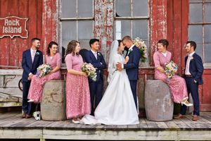 Wedding party poses outside rustic Rock Island barn, bride and groom kissing flanked by bridesmaids in pink and groomsmen.