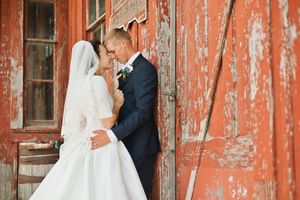 A bride and groom share a romantic moment leaning against a rustic red barn door on their wedding day.