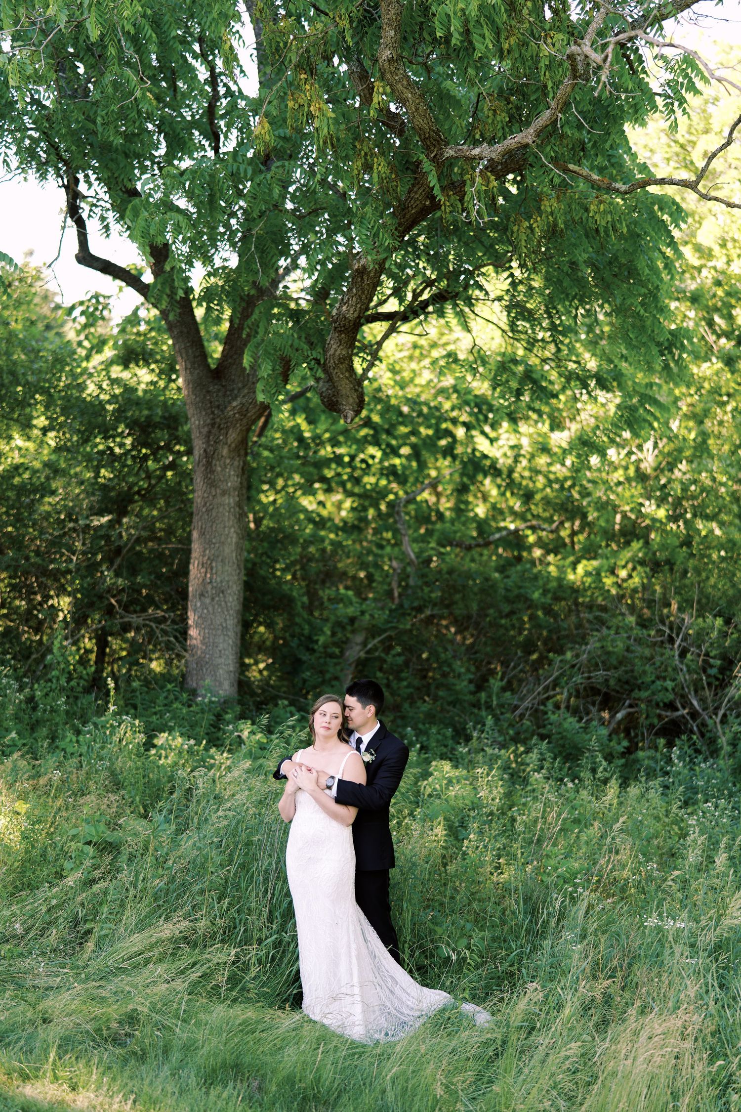 bride and groom embracing in a sunny summer scene with long grass in the foreground and tall trees in the background