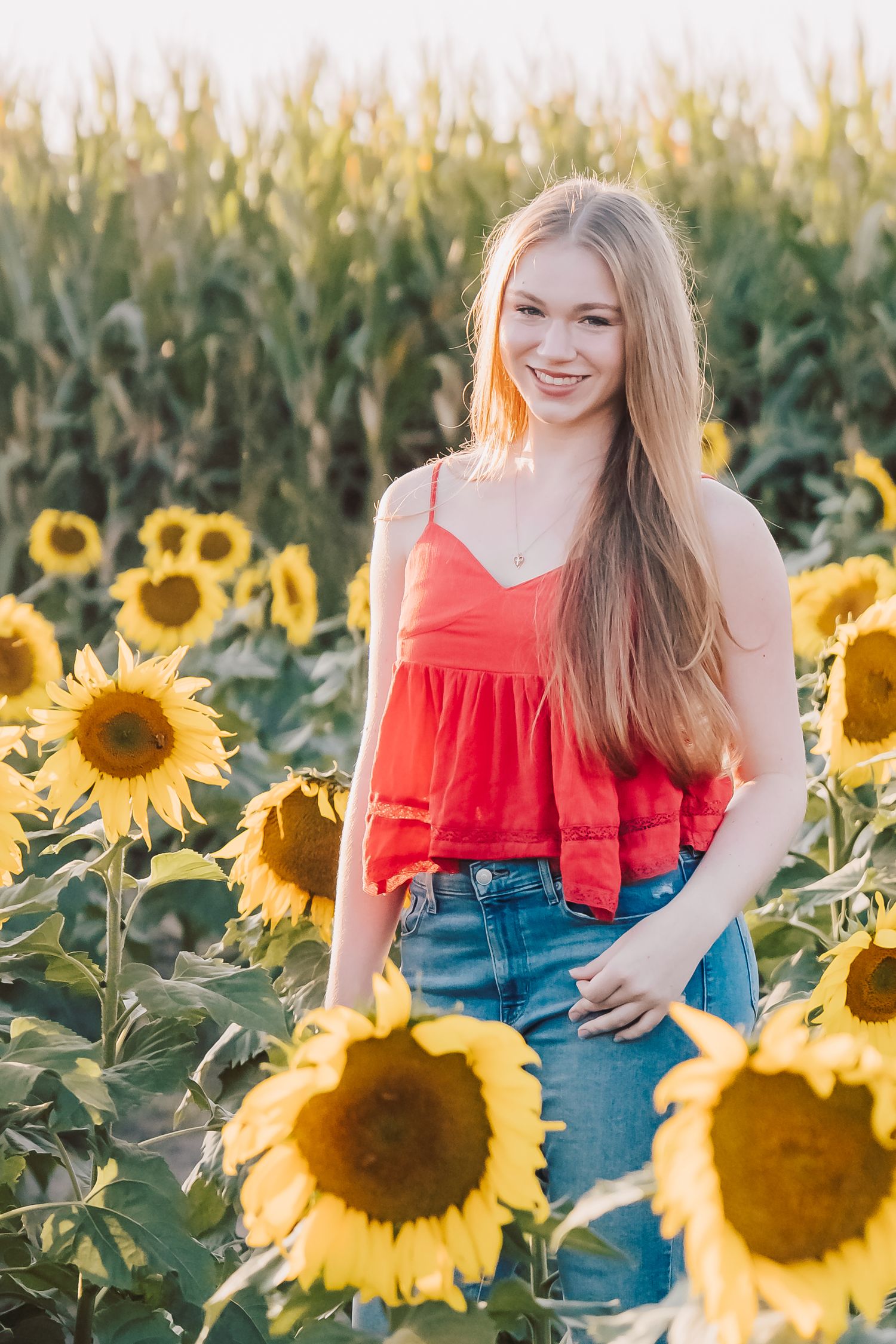 A person in a red top and jeans poses cheerfully among bright yellow sunflowers in a summer field at golden hour.