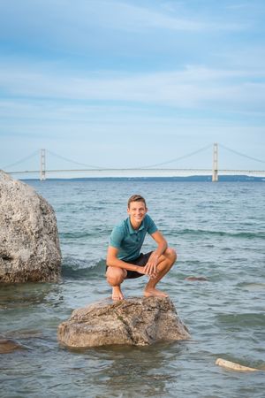 Person poses on rocks in the water with Golden Gate Bridge visible in background across San Francisco Bay.