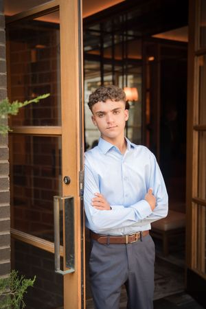 A young professional in a light blue dress shirt and gray slacks stands confidently in a doorway of a modern building.