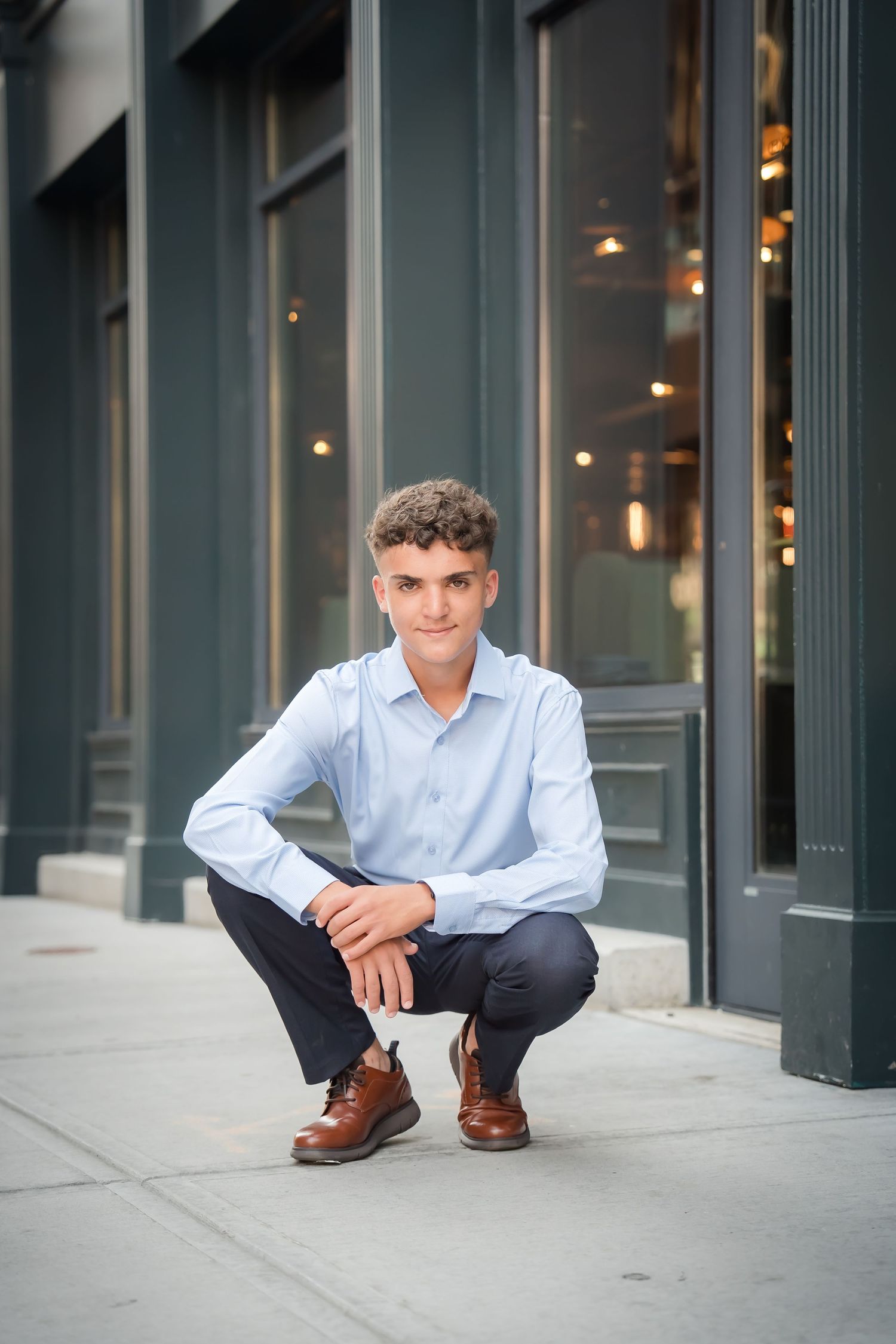 A person crouches on a sidewalk wearing a light blue button-down shirt, dark pants and brown leather shoes near a storefront.