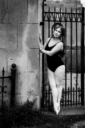 A ballet dancer in black leotard poses en pointe by an ornate iron gate.