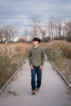 Person in green hoodie walking on wooden boardwalk through tall grass.