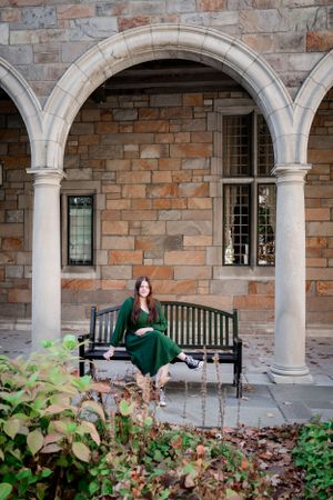 Person in emerald dress sitting on bench under stone archways of historic building with foliage.